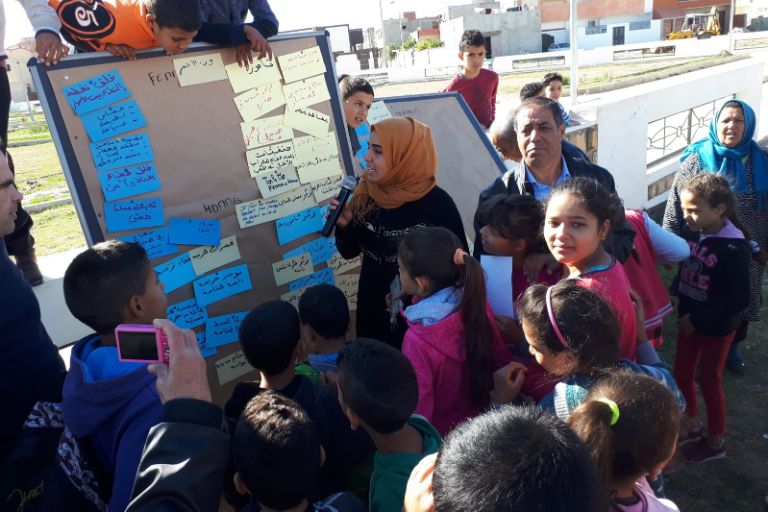 Eine Frau mit Mikrofon und viele Kinder stehen vor einer Pinnwand in Kasserine und präsentieren die Ergebnisse der Planung