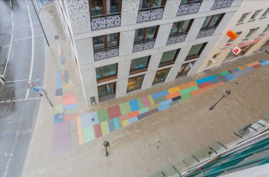 Bodenmosaik mit unterschiedlich großen farbige Steinen, der sich um die Ecke des Gebäudes am Eck Oberanger / Dultstraße legt. In zwei der Bodenplatten ist ein rosa und ein schwarzes Dreieck eingelassen.