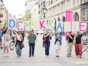 Auf dem Bild sind sieben Jugendliche zu sehen. Sie gehen durch die Sendlinger Straße und halten die Buchstaben des Wortes Demokratie in die Luft.