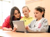 Das Foto zeigt zwei Grundschüler*innen mit einem Tablet in der Hand und eine Lehrkraft, die mit beiden Inhalte darauf ansieht. 