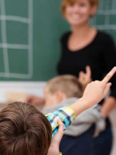 Lehrerin steht an der Tafel und Kinder melden sich.
